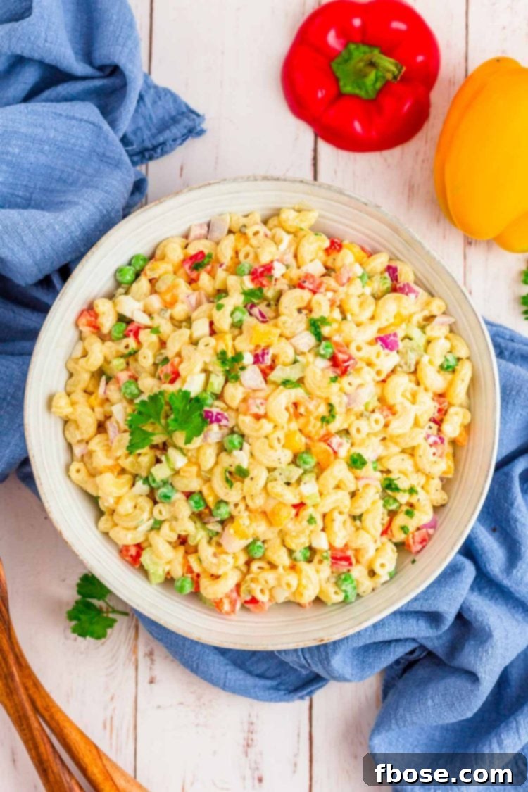 An overhead view of a large, rustic wooden bowl filled with creamy macaroni salad, garnished with fresh herbs, sitting on a pristine white wooden table. The salad is rich in tender macaroni and colorful diced vegetables.