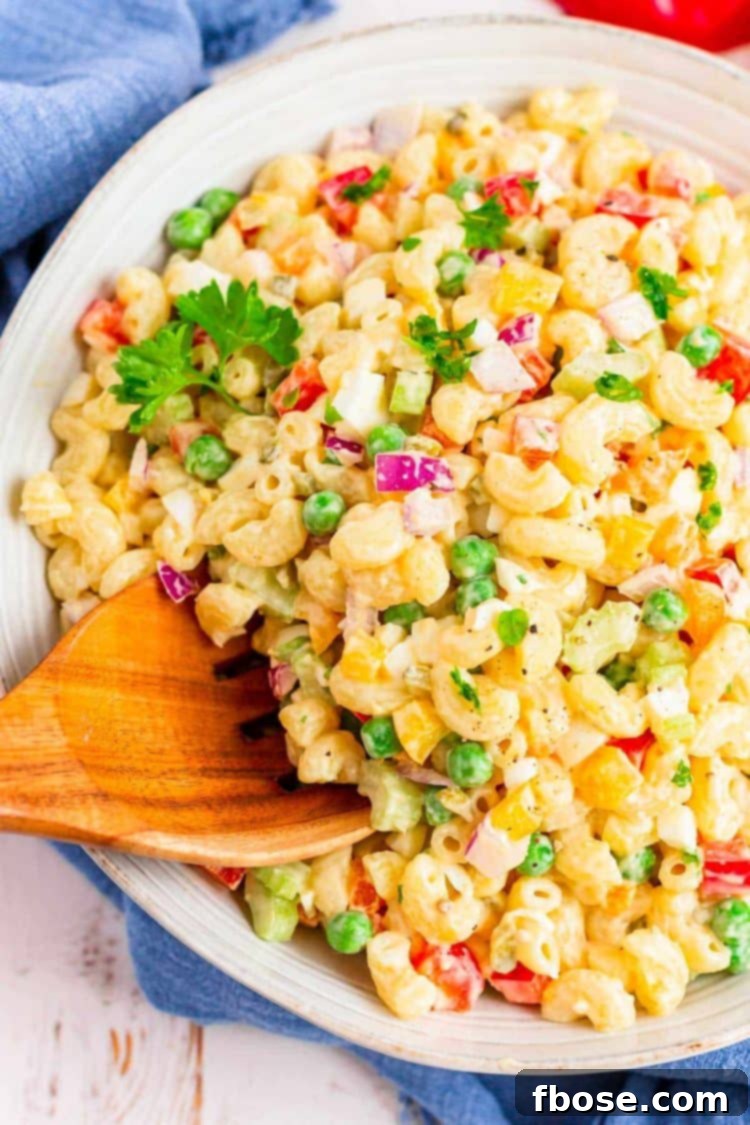 A wooden serving spoon scooping a generous portion of classic macaroni salad from a large white bowl, showcasing the creamy dressing, tender macaroni, and colorful vegetables.