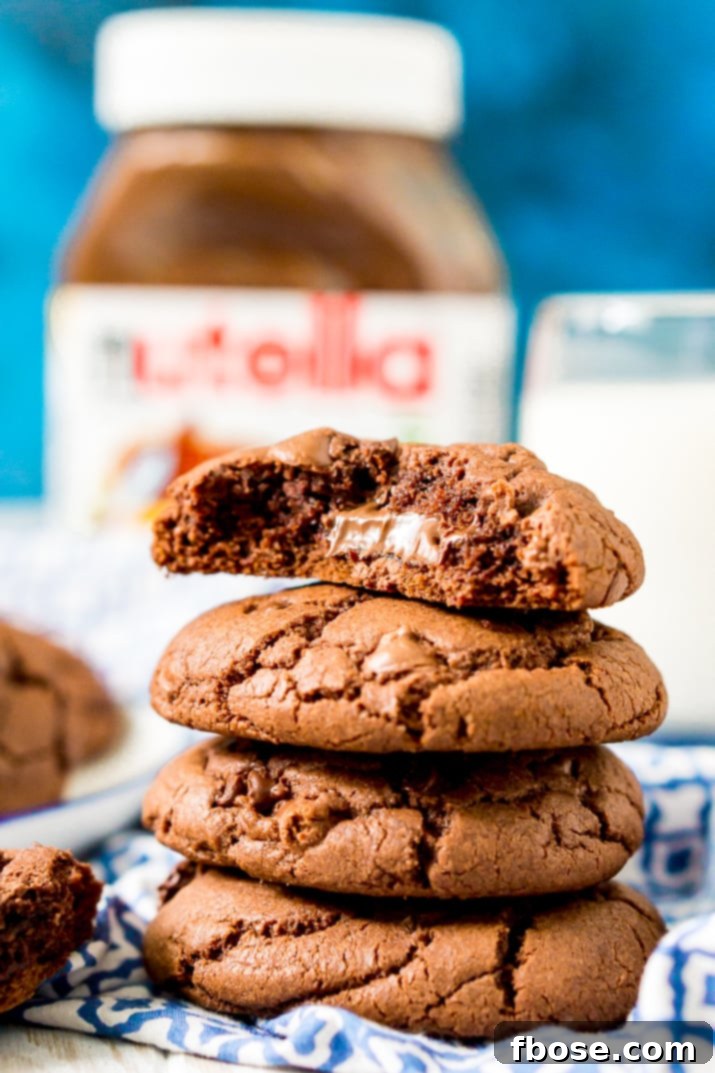 Gooey Nutella Stuffed Cookies 4 Close up photo of a stack of 4 chocolate cookies on a blue and white napkin, the top one has a bite taken out of it revealing nutella.