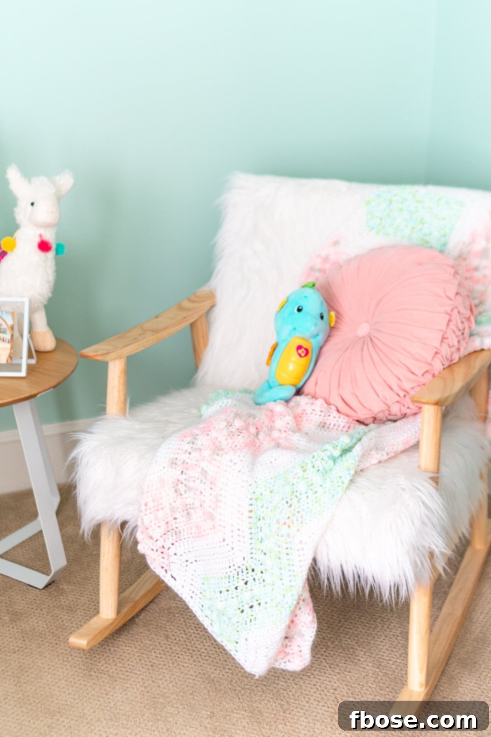 A cozy faux sheepskin and wooden rocking chair, adorned with a knitted blanket and pink pillow, creating a warm corner in the baby nursery.
