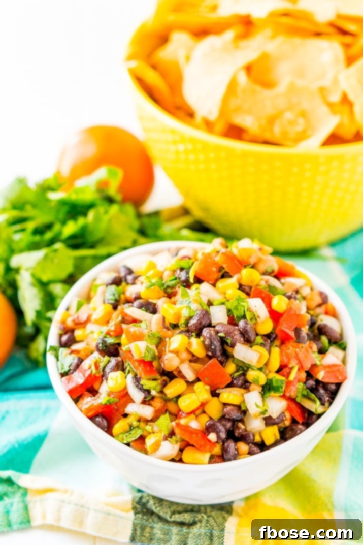 A large bowl of colorful Cowboy Caviar sits on a teal and yellow napkin, with a smaller bowl of tortilla chips in the background, inviting guests to dip and enjoy this fresh and hearty Mexican-inspired appetizer.