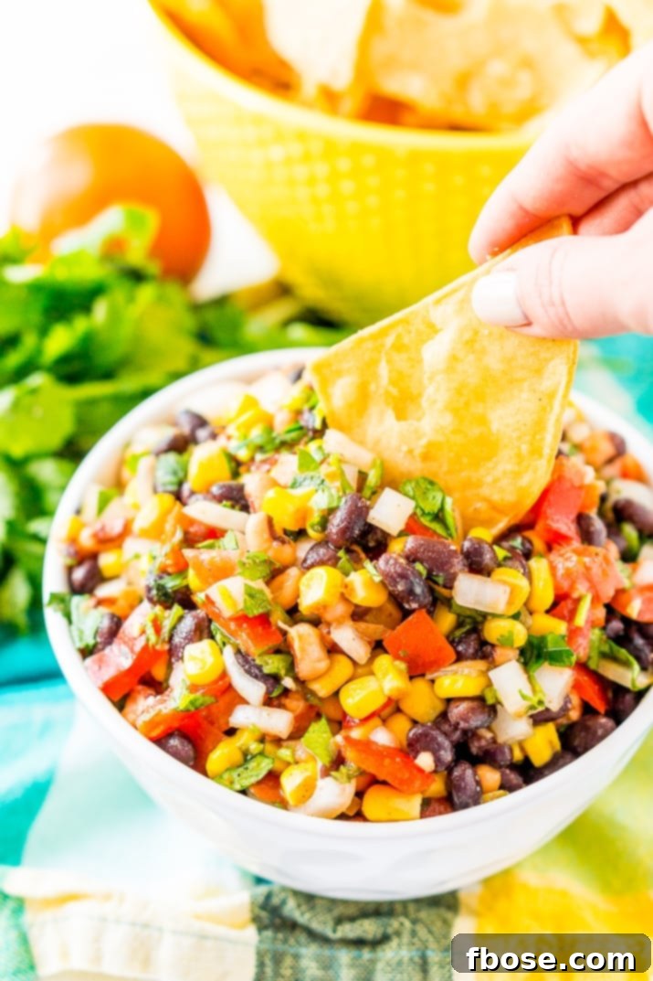 A woman's hand gracefully dips a crisp tortilla chip into a colorful bowl of Cowboy Caviar, highlighting the perfect scooping consistency and inviting texture of this popular Mexican-inspired dip.