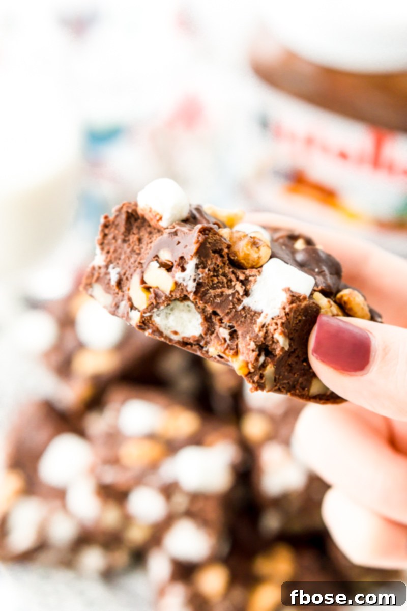 A hand reaching for a piece of Nutella Rocky Road Fudge from a white plate, highlighting the irresistible nature of the treat, rich with marshmallows, walnuts, and chocolate chips.