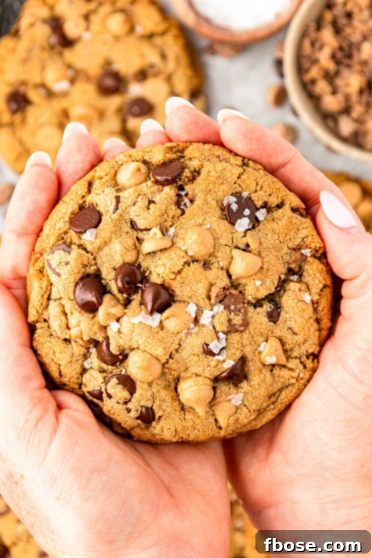 A woman's hand holding a chocolate chip cookie with oatmeal, peanut butter chips, and toffee in it.