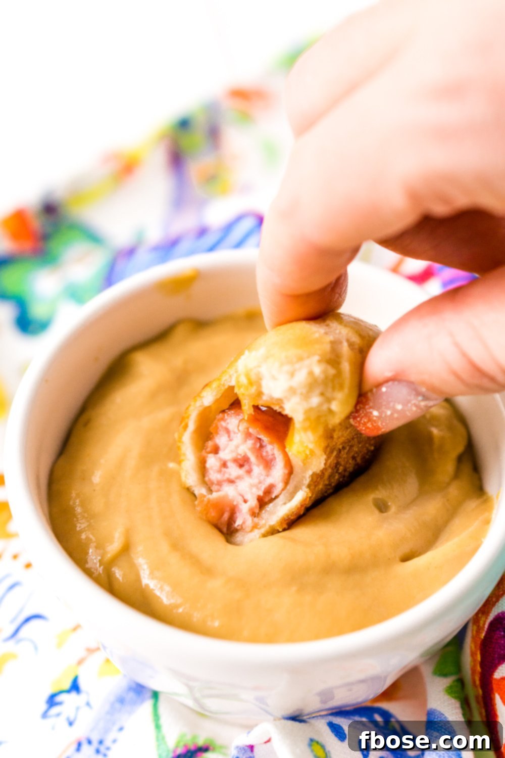 Woman's hand dipping pigs in a blanket into dipping sauce with bite taken out of it.