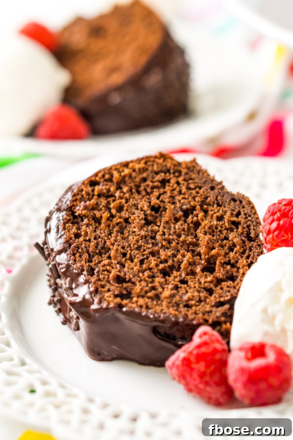A tempting slice of Healthier Chocolate Yogurt Cake on a white plate, served with a scoop of ice cream and fresh raspberries, highlighting its moist texture.