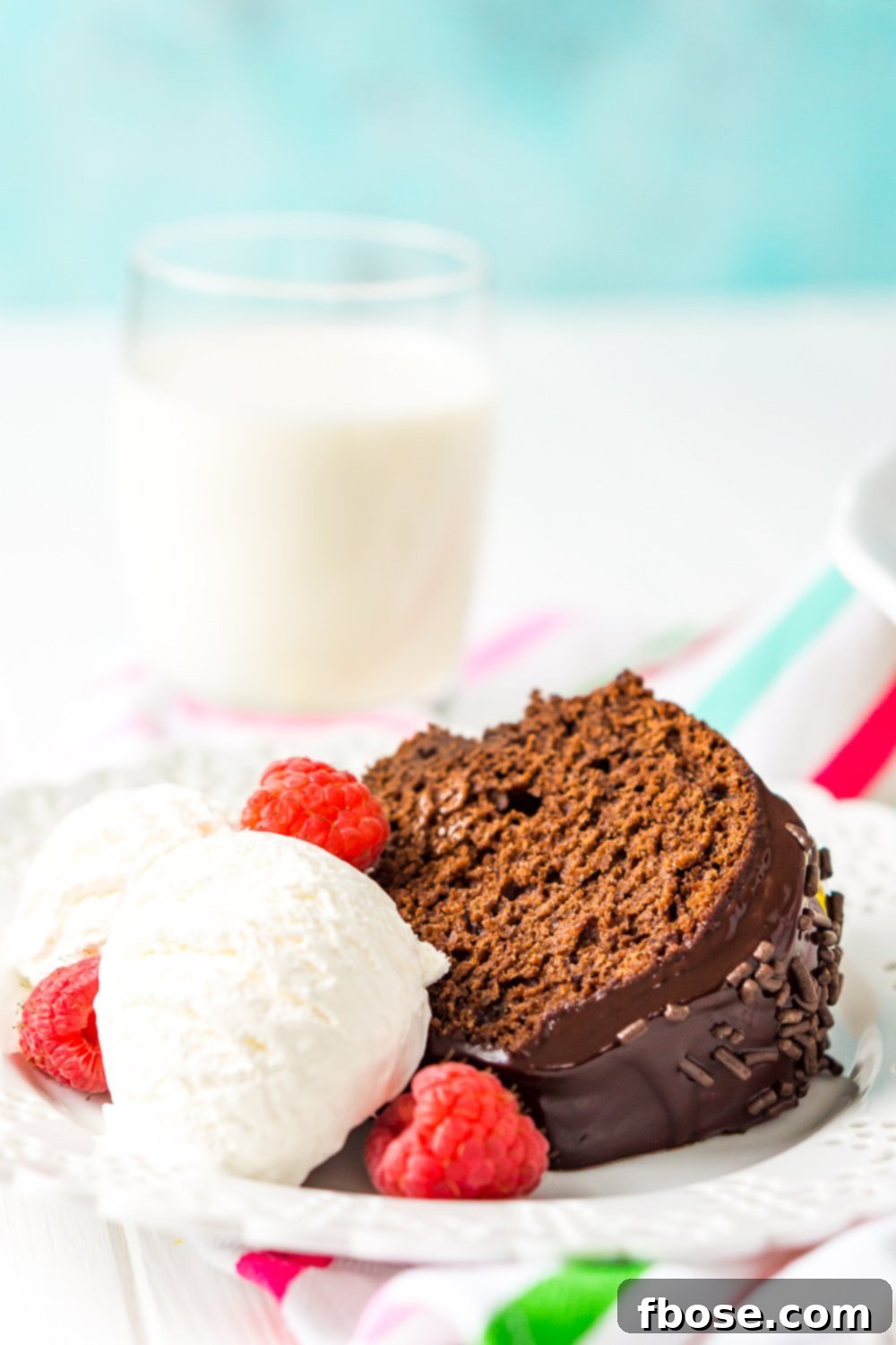 A delicious slice of healthy chocolate cake on a white plate, accompanied by a scoop of ice cream and fresh raspberries, served with a refreshing glass of milk.