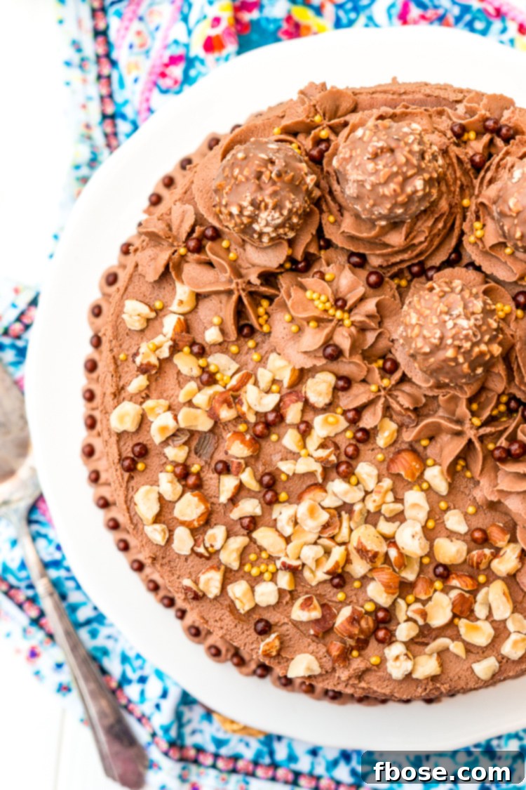 Close-up photo of the top of a decadent Nutella chocolate cake, garnished with chopped hazelnuts and whole Ferrero Rocher candies.