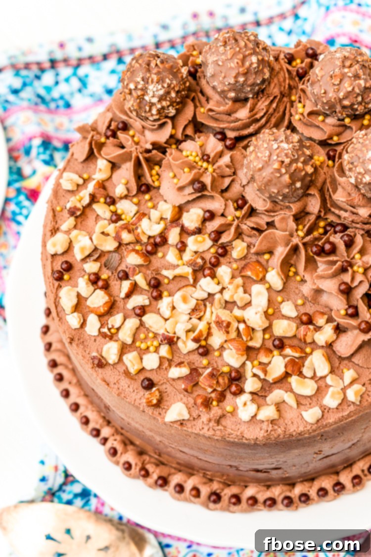 A stunning Nutella hazelnut cake on a white cake stand, beautifully decorated with Ferrero Rocher candies and chopped hazelnuts.