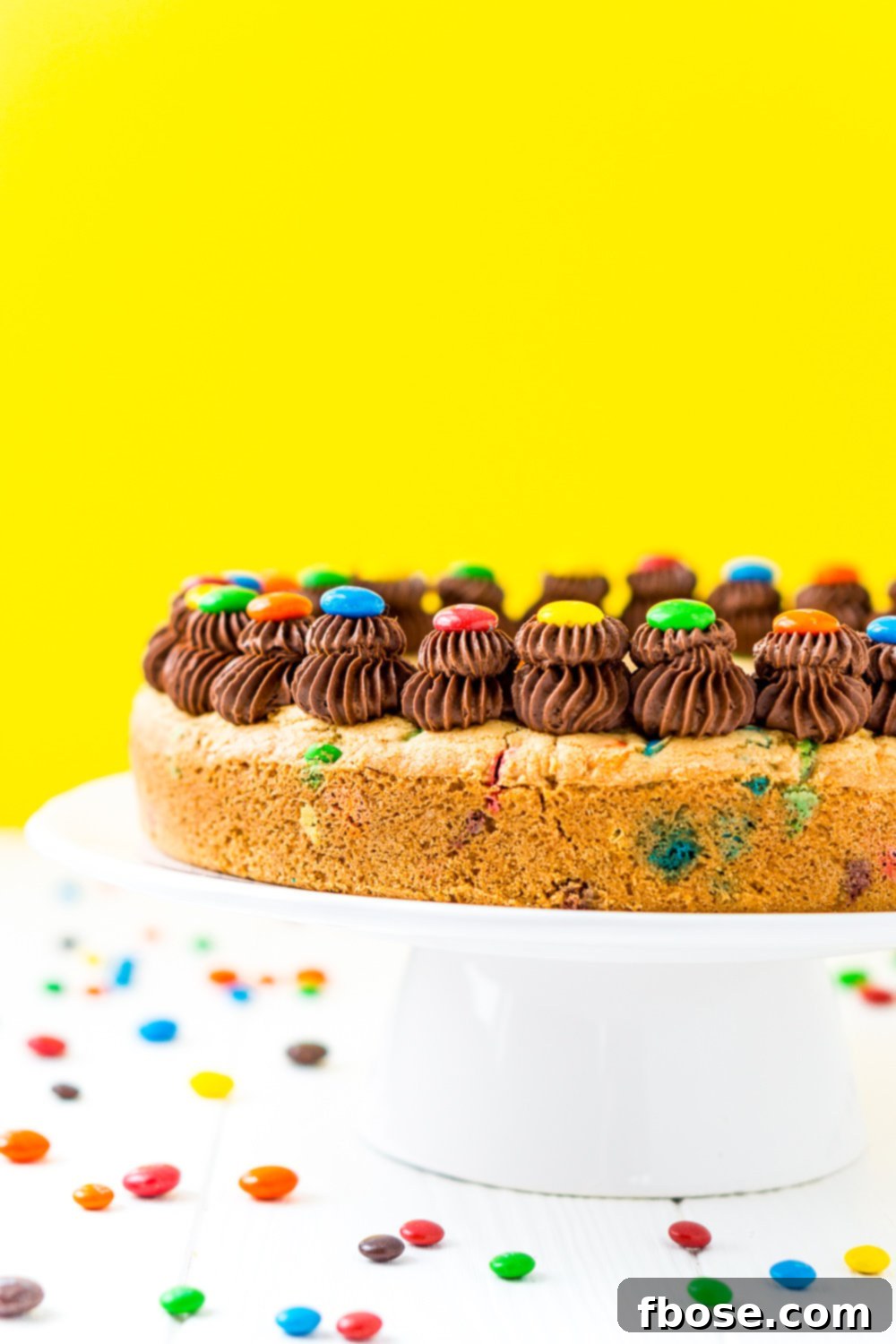 M&M's Cookie Cake with chocolate frosting on a white cake stand with yellow background.