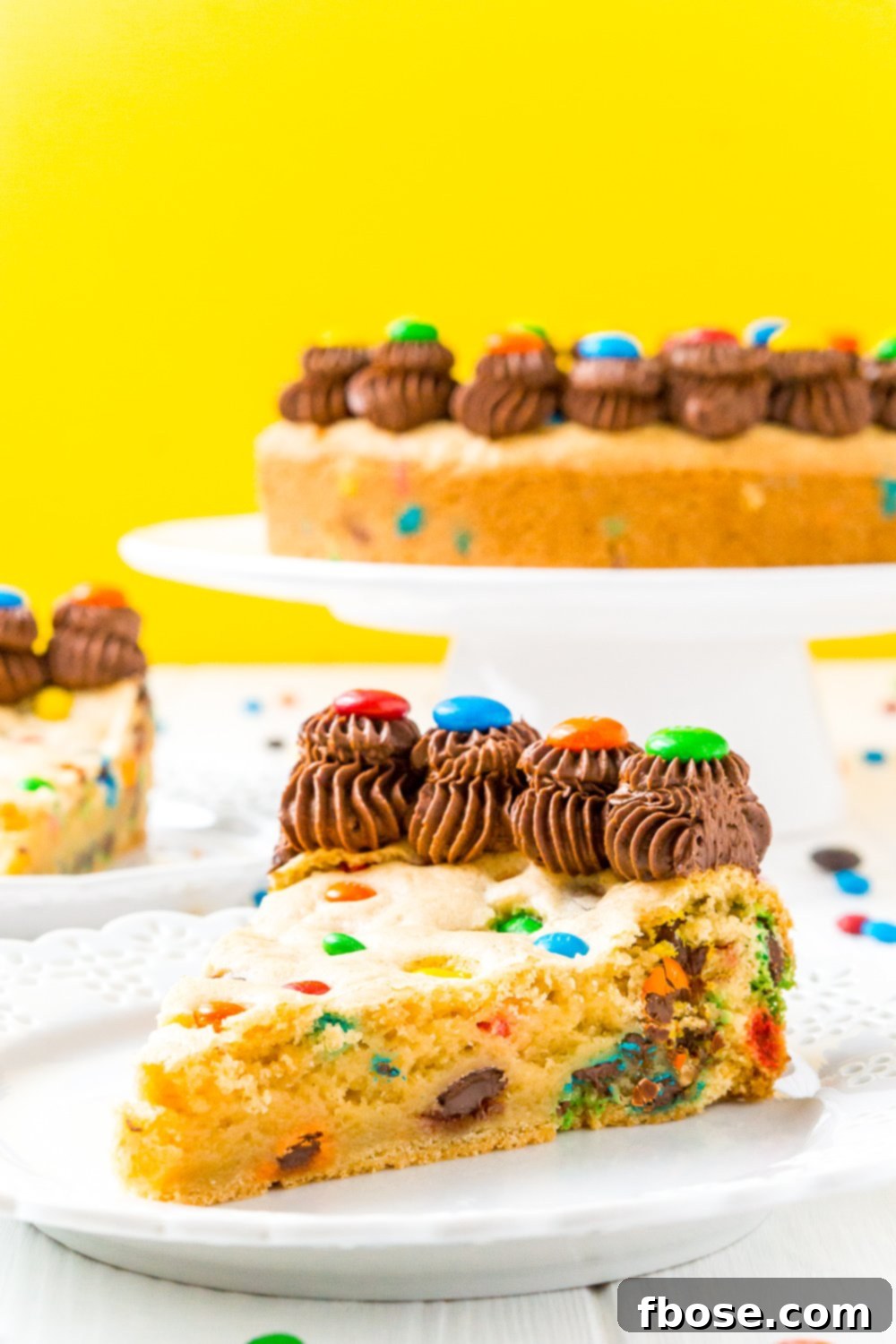 Slice of M&M's Cookie Cake on white plate with rest of cake in the background.