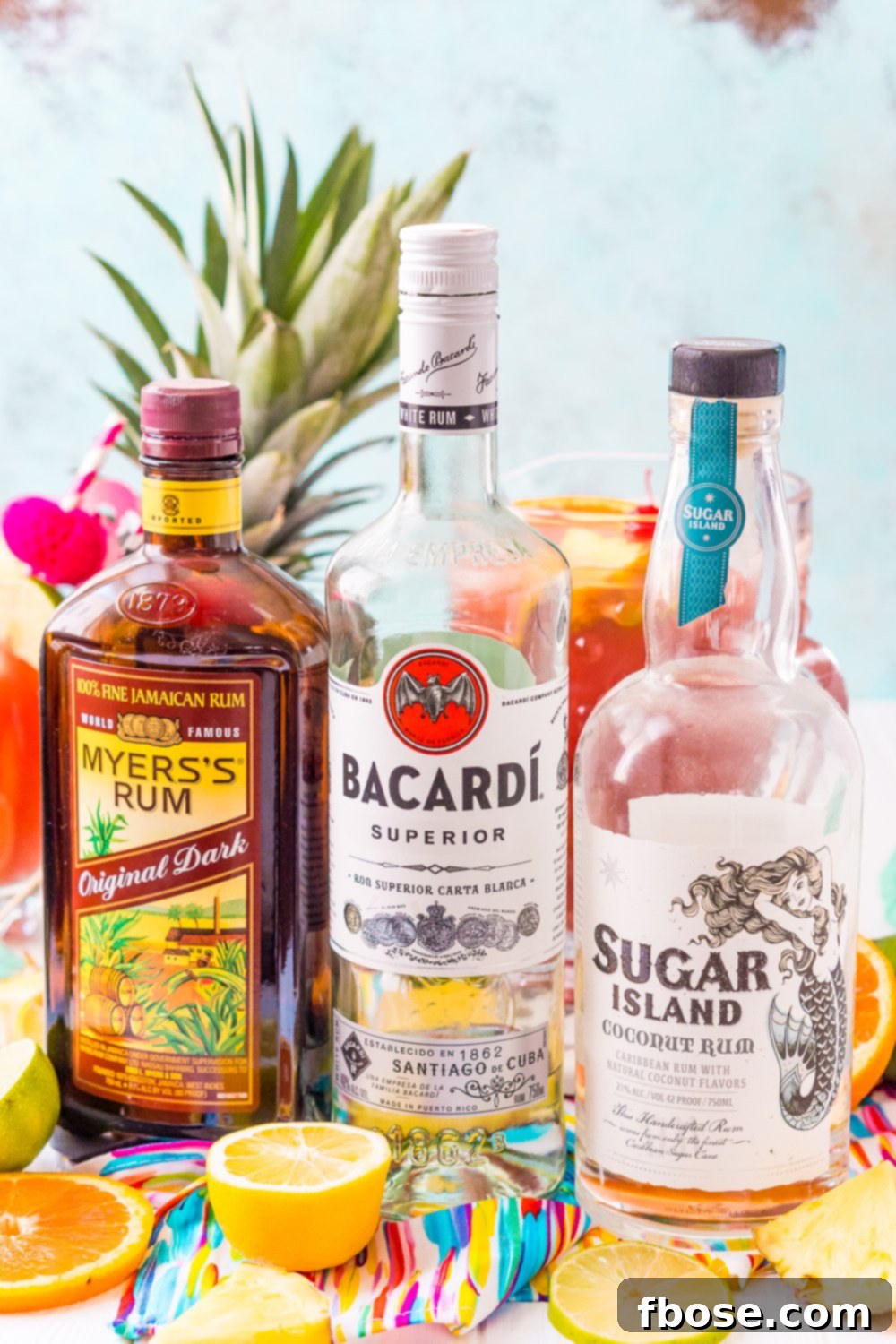 Bottles of Myers's Dark Rum, Bacardi White Rum, and Sugar Island Coconut Rum are displayed alongside a pitcher of freshly made Rum Punch and a whole pineapple, highlighting the key ingredients for this delicious Caribbean cocktail.