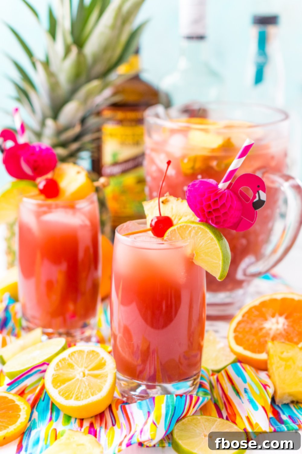 Multiple glasses of Rum Punch, each adorned with a cherry, pineapple, and lime, accompanied by festive flamingo paper straws and a pitcher in the background, showcasing an inviting party spread.