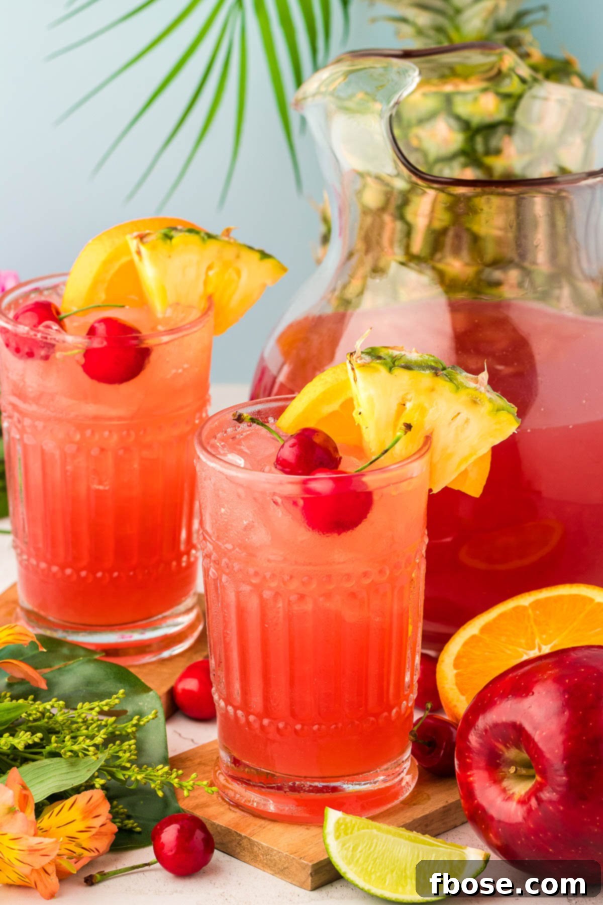 A glass of fruit punch on a wooden coaster with another glass, pitcher, and fruit in the background.