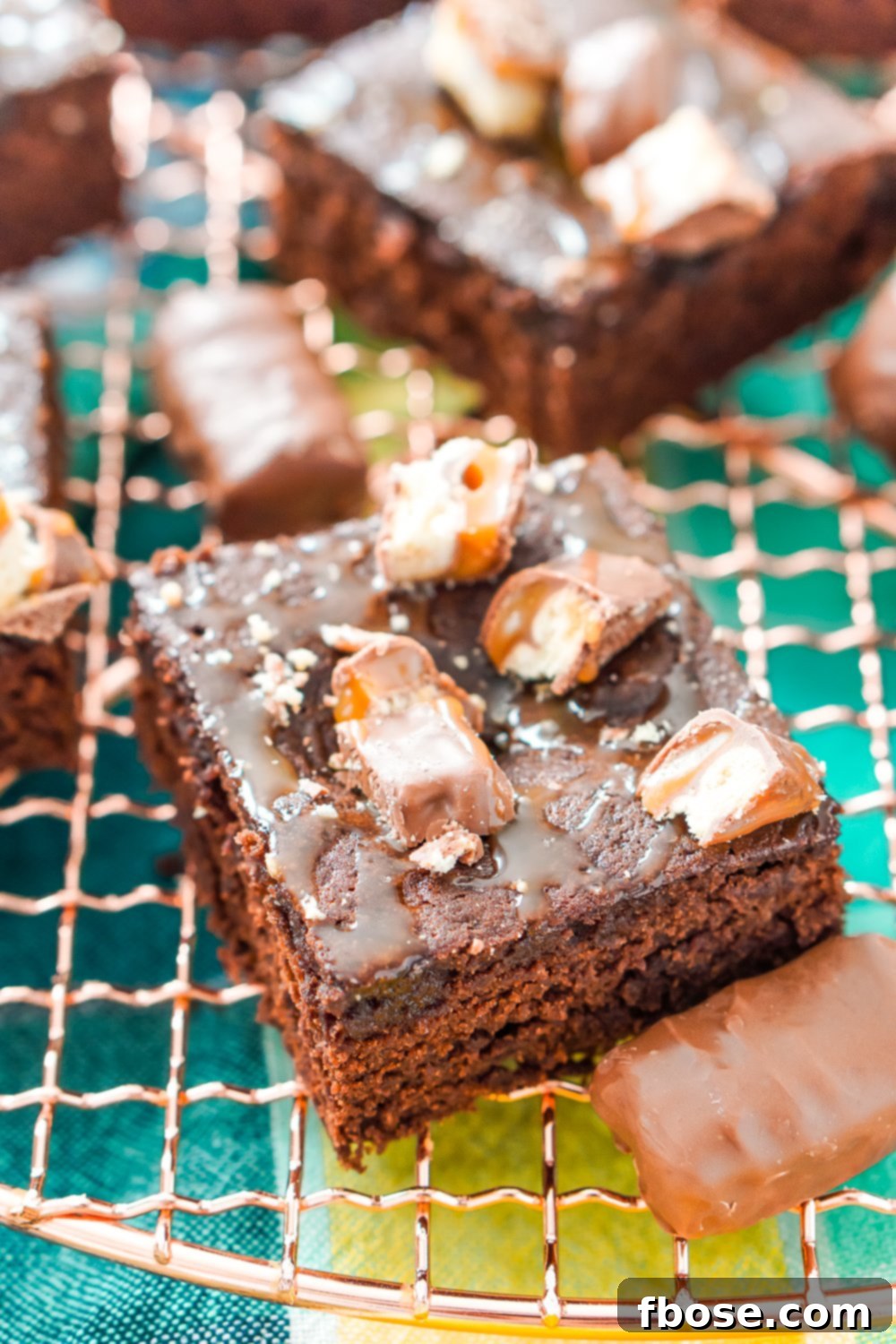 Twix Caramel Fudge Brownies 3 A square Twix Caramel Brownie on a copper cooling rack with more brownies and mini Twix bars surrounding it.