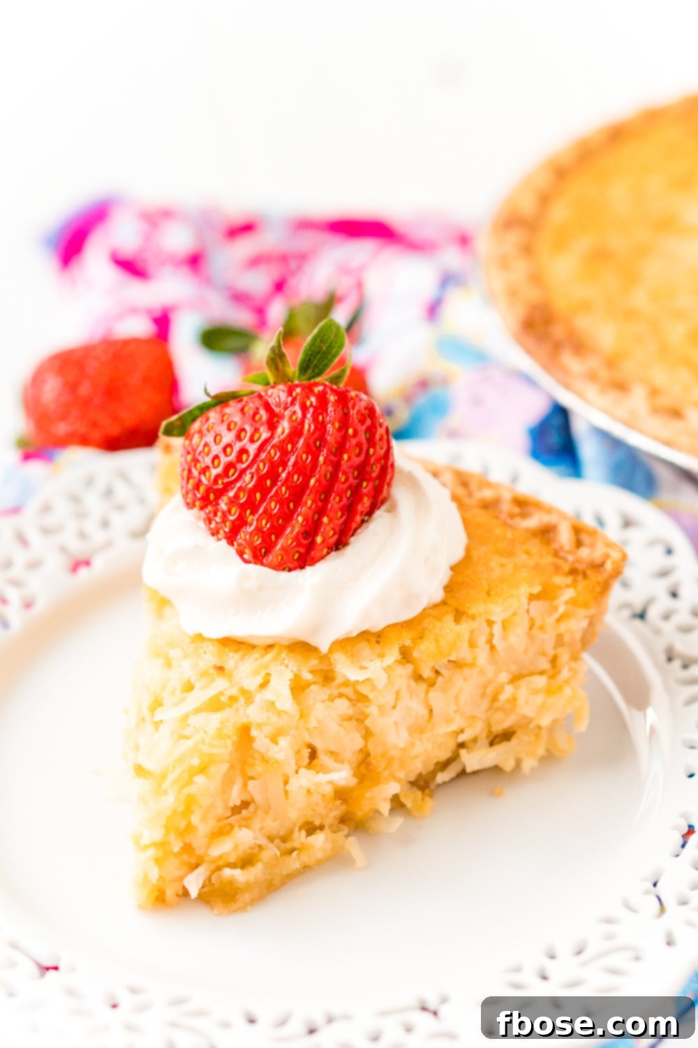 Slice of Coconut Custard Pie on a white plate with whipped cream and a strawberry on top.