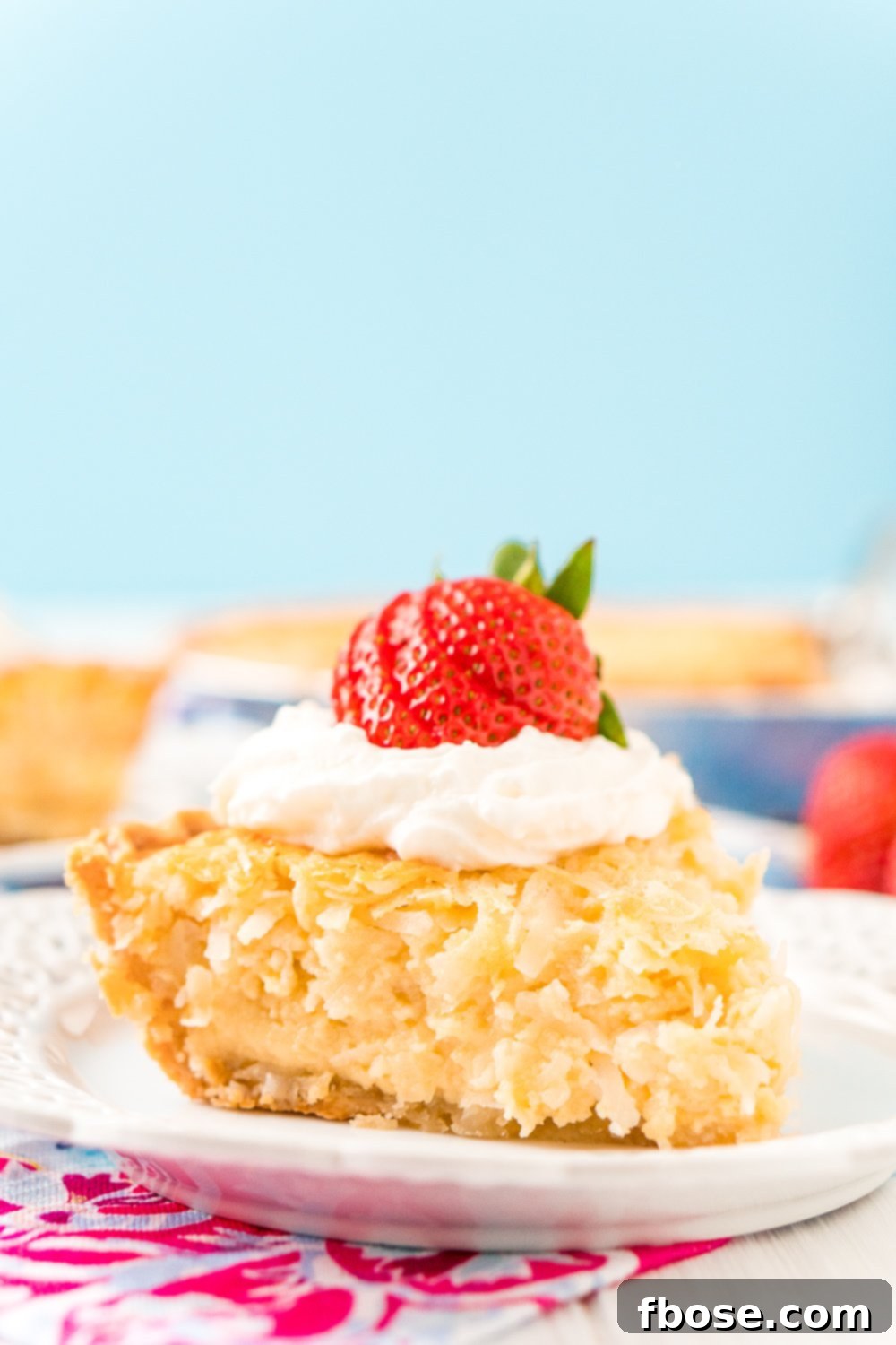 Slice of Coconut Custard Pie on a white plate with pink and white napkin and a blue background. Pie has whipped cream and a strawberry on top.