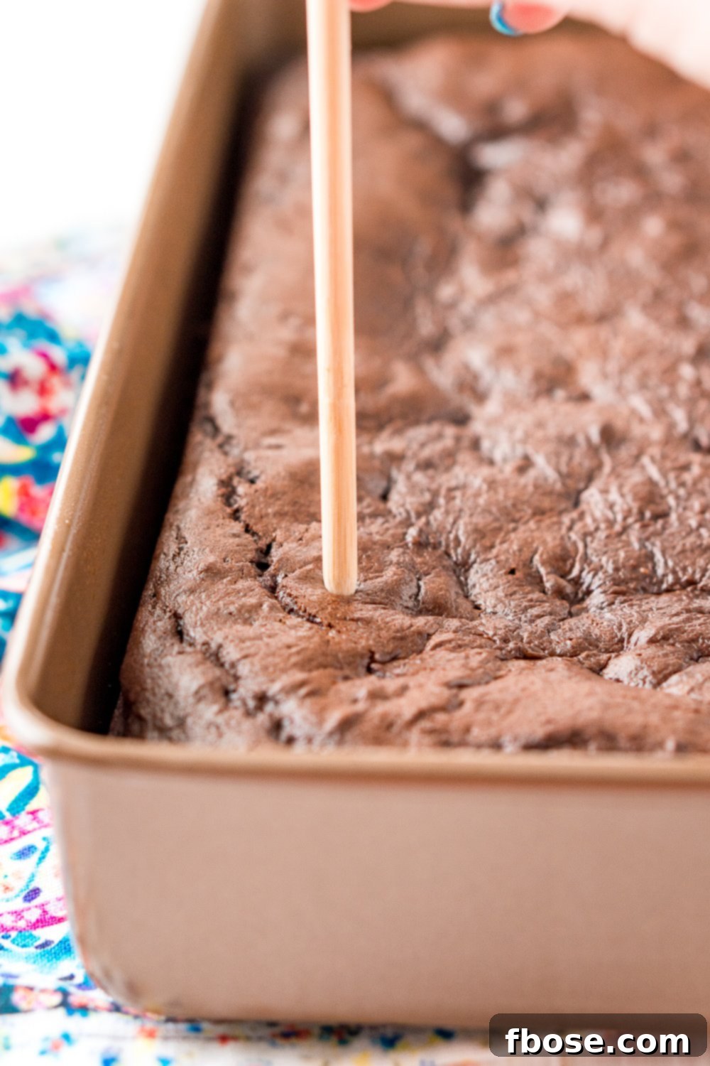 Chocolate cake in a 9x13-inch pan with a woman's hand holding a wooden skewer poking holes in the cake.