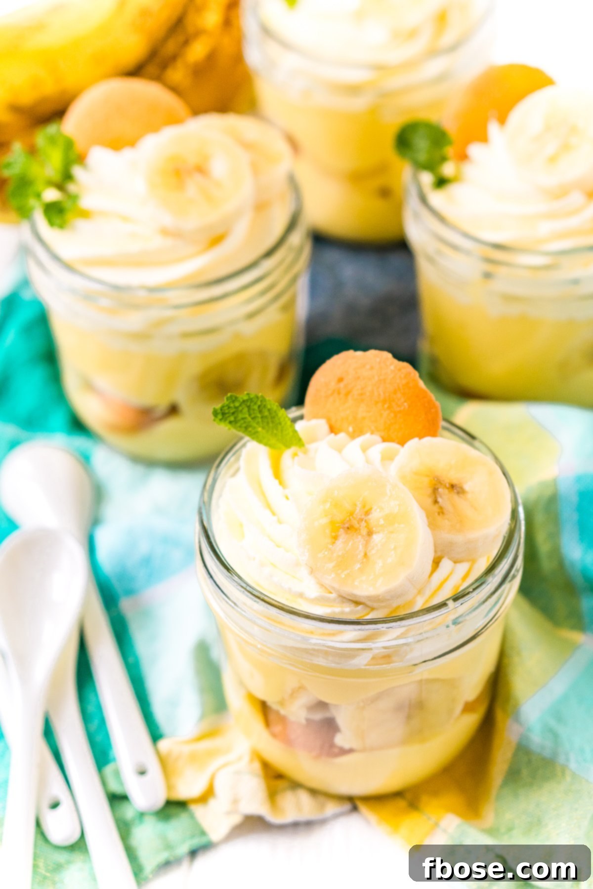 Mini mason jars with banana pudding in them on a blue and yellow napkin with white spoons to the side.