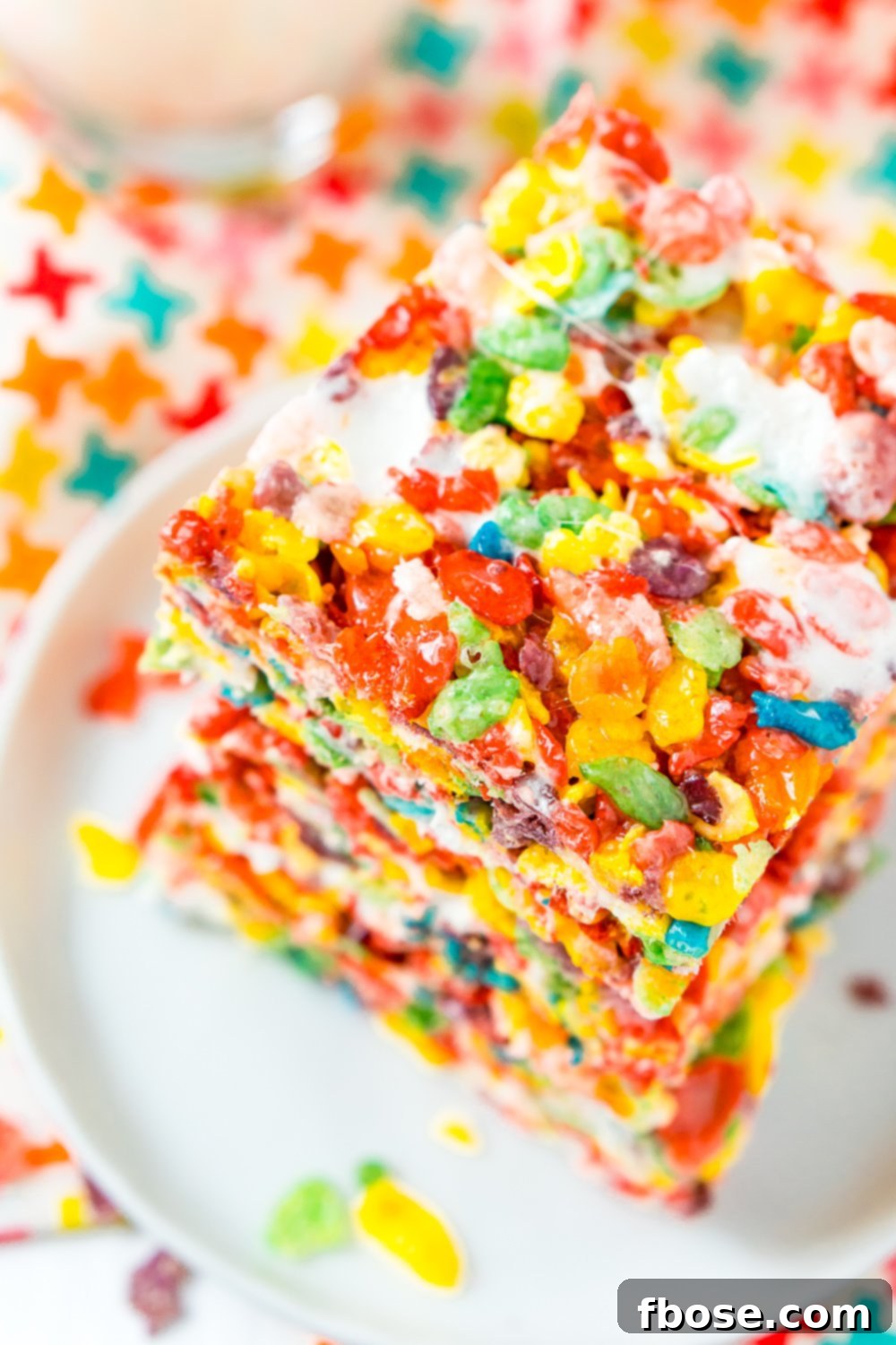 A stack of colorful Fruity Pebbles Treats sits invitingly on a small white plate, placed on a vibrant, patterned napkin. The soft lighting highlights their unique texture and appealing appearance.