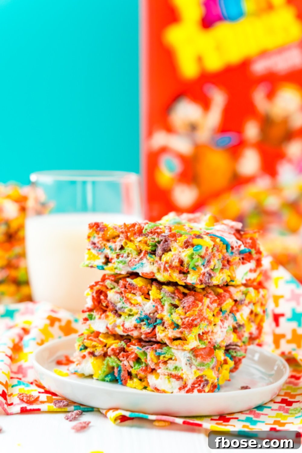 A charming scene featuring a stack of Fruity Pebbles Treats on a white plate, accompanied by a refreshing glass of milk and a box of Fruity Pebbles cereal in the background. A perfect morning or afternoon snack.