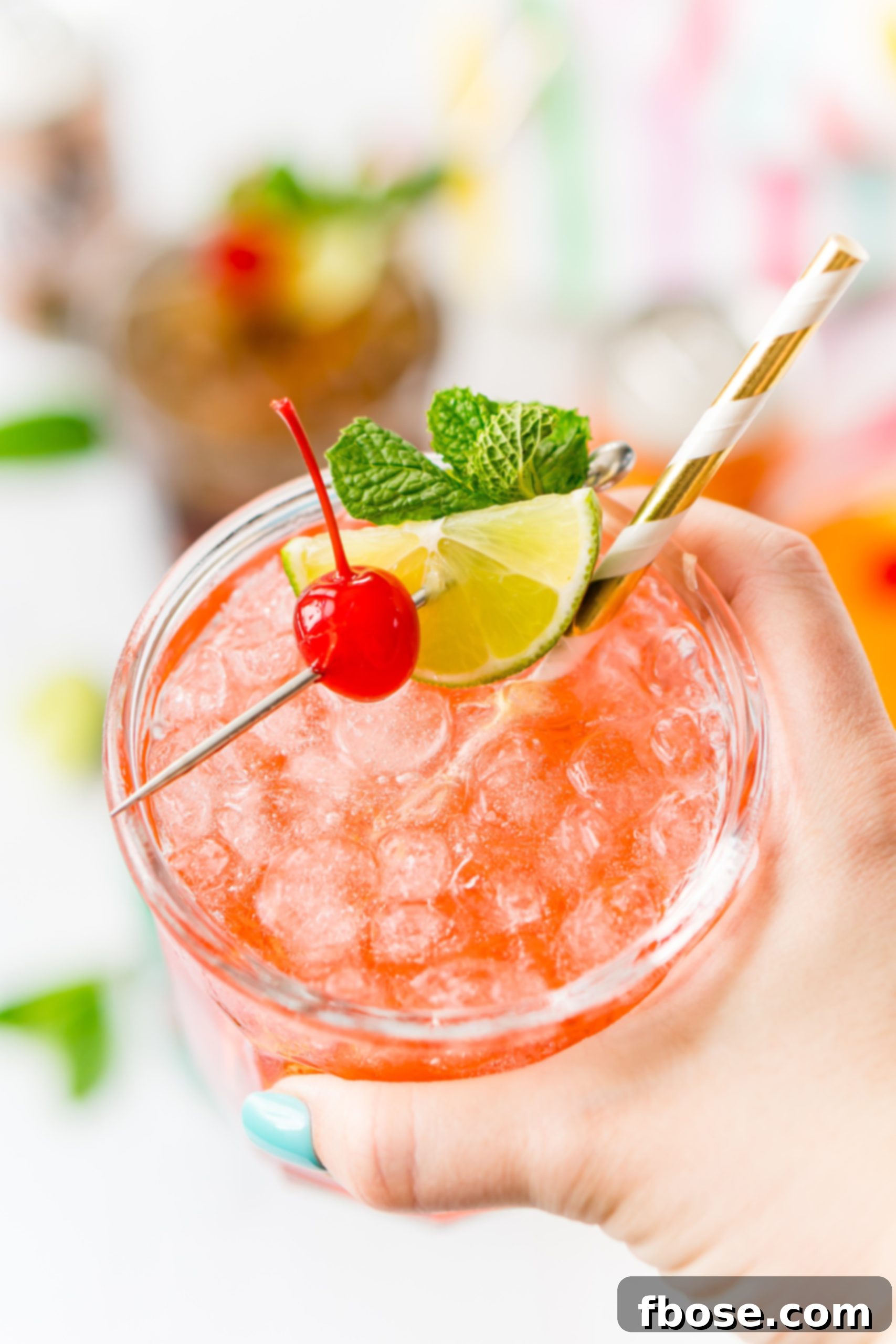 Simple Sips Three Ways 6 A woman's hand elegantly holding up a vibrant mocktail glass, garnished with a bright red cherry, a fresh mint sprig, and a crisp lime wedge, showcasing the drink's appealing presentation.