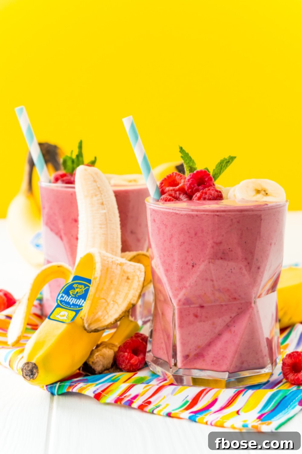 Berry Banana Bliss 3 Straight on photo of two glasses filled with raspberry banana smoothie with a banana leaning next to the glass.