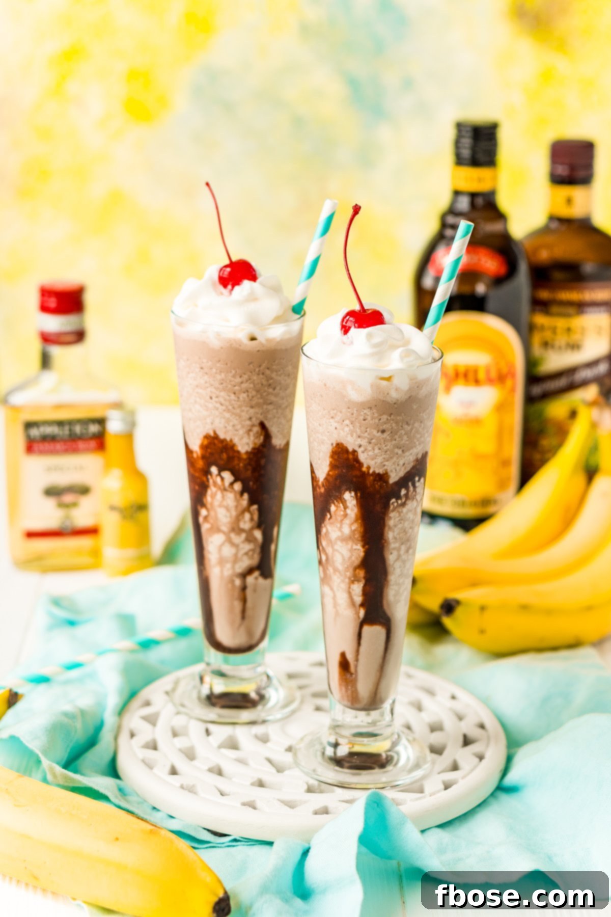 Two dirty banana drinks on a white wooden board with bottles of rum and bananas in the background.