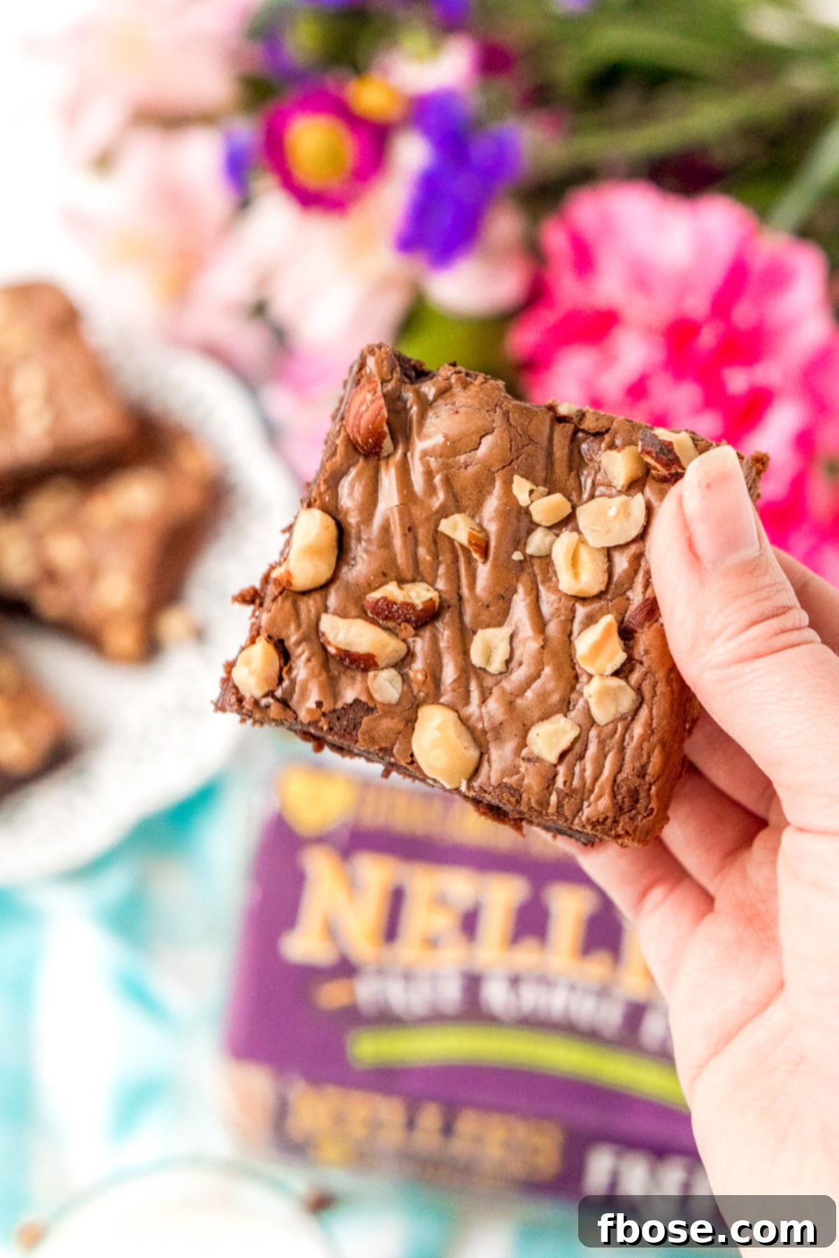 Woman's hand holding a hazelnut brownie. More brownies and flowers in the background.