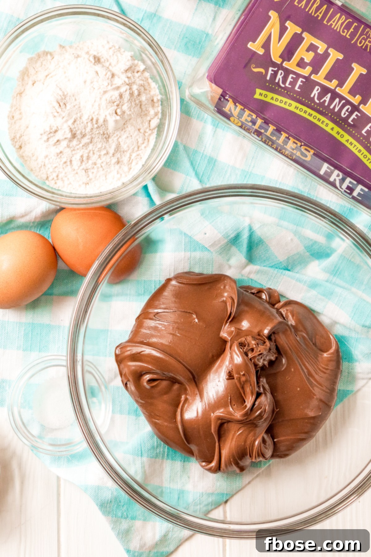 Nutella in a glass bowl with carton of eggs, loose eggs, small bowl of flour and salt on a white and blue napkin.