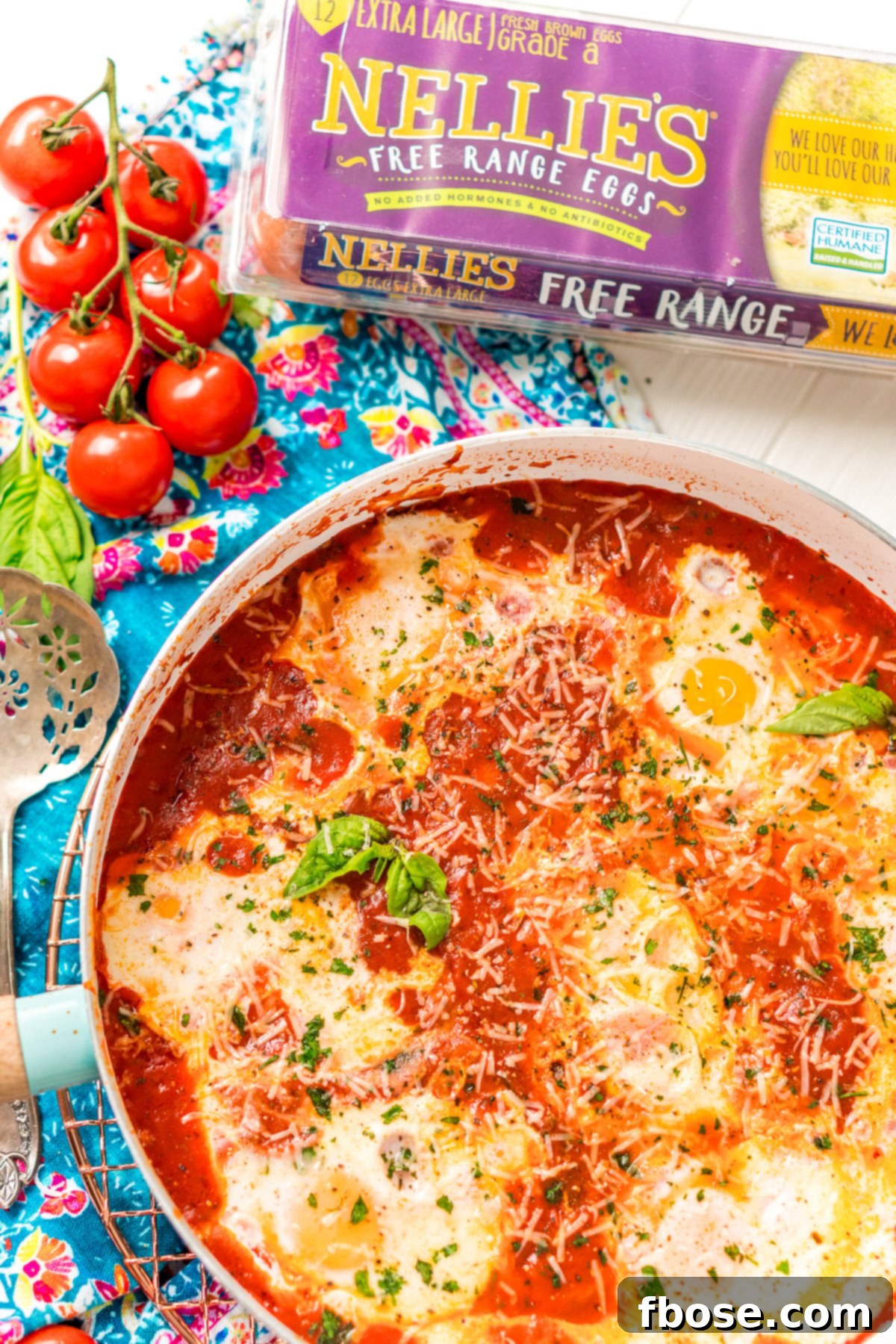 Eggs In Purgatory in a white and blue skillet with a blue napkin. Tomatoes and a carton of eggs are to the side of the pan. A beautifully presented skillet of Eggs in Purgatory, featuring bright red tomato sauce and creamy poached eggs, ready to be served.