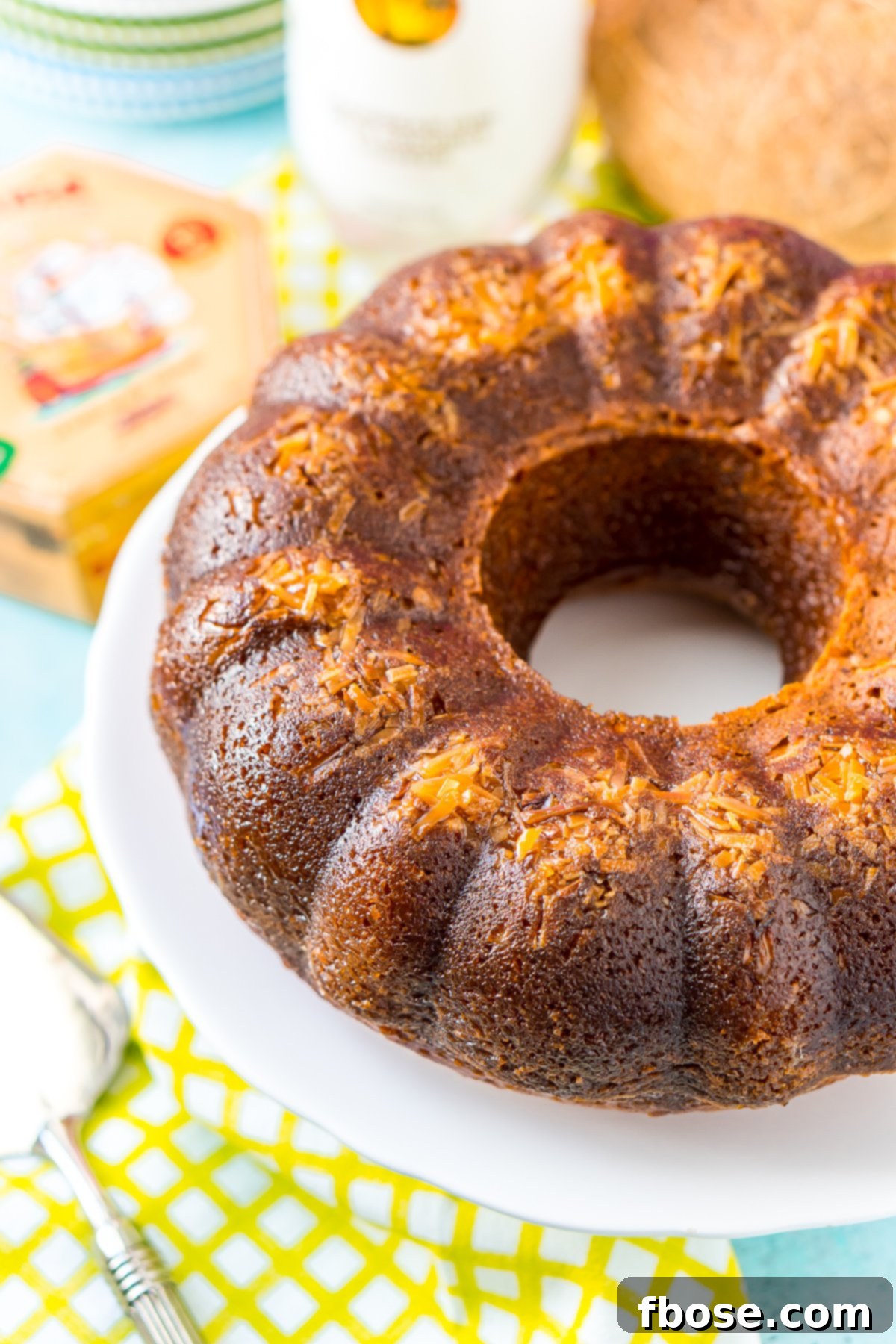 Caribbean Coconut Rum Bliss 3 Whole bundt cake with coconut baked into the top on a white cake stand with a bottle of rum in the background.