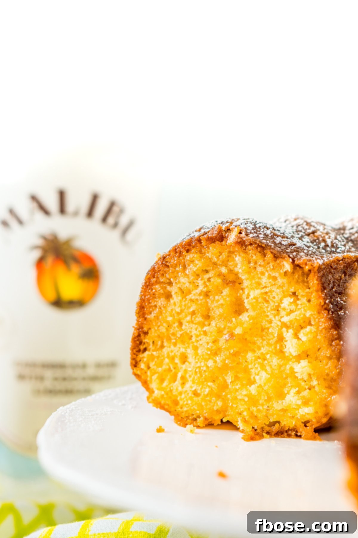 Caribbean Coconut Rum Bliss 5 Rum cake with a slice taken out of it on a cake stand with a bottle of coconut rum in the background.