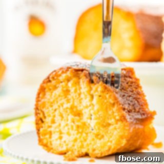Fork taking a bite out of a slice of coconut rum cake on a white plate with a bottle of rum and more cake in the background.