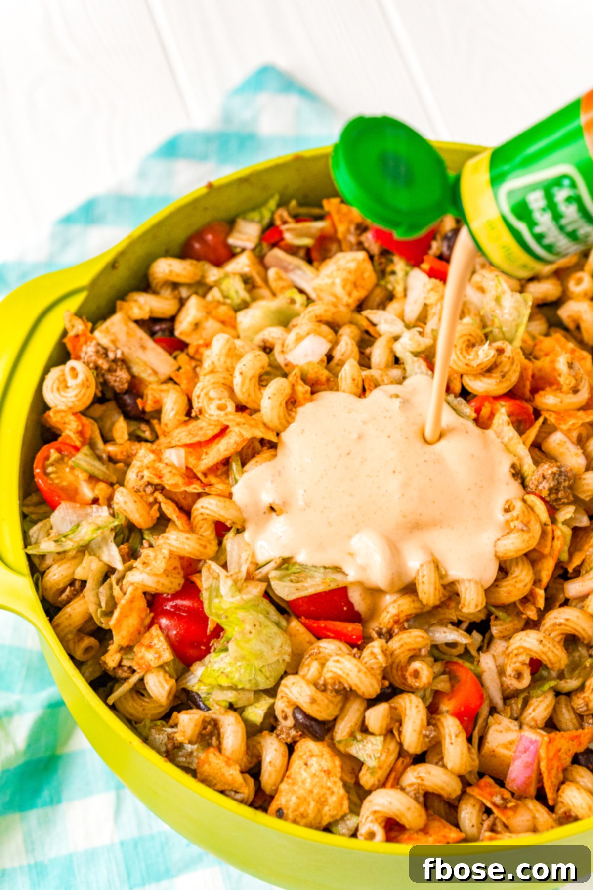 A large bowl of taco-flavored pasta salad being generously drizzled with creamy dressing, highlighting the rich and flavorful sauce before tossing.