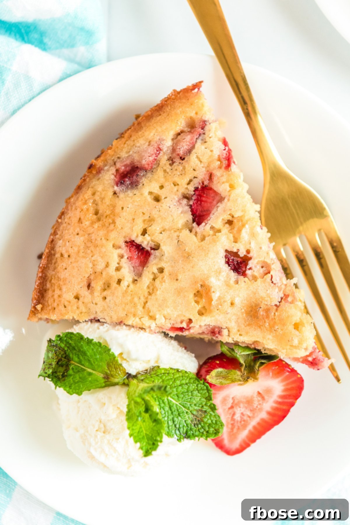 Overhead photo of a slice of strawberry cake on a white plate with whipped cream and gold fork.