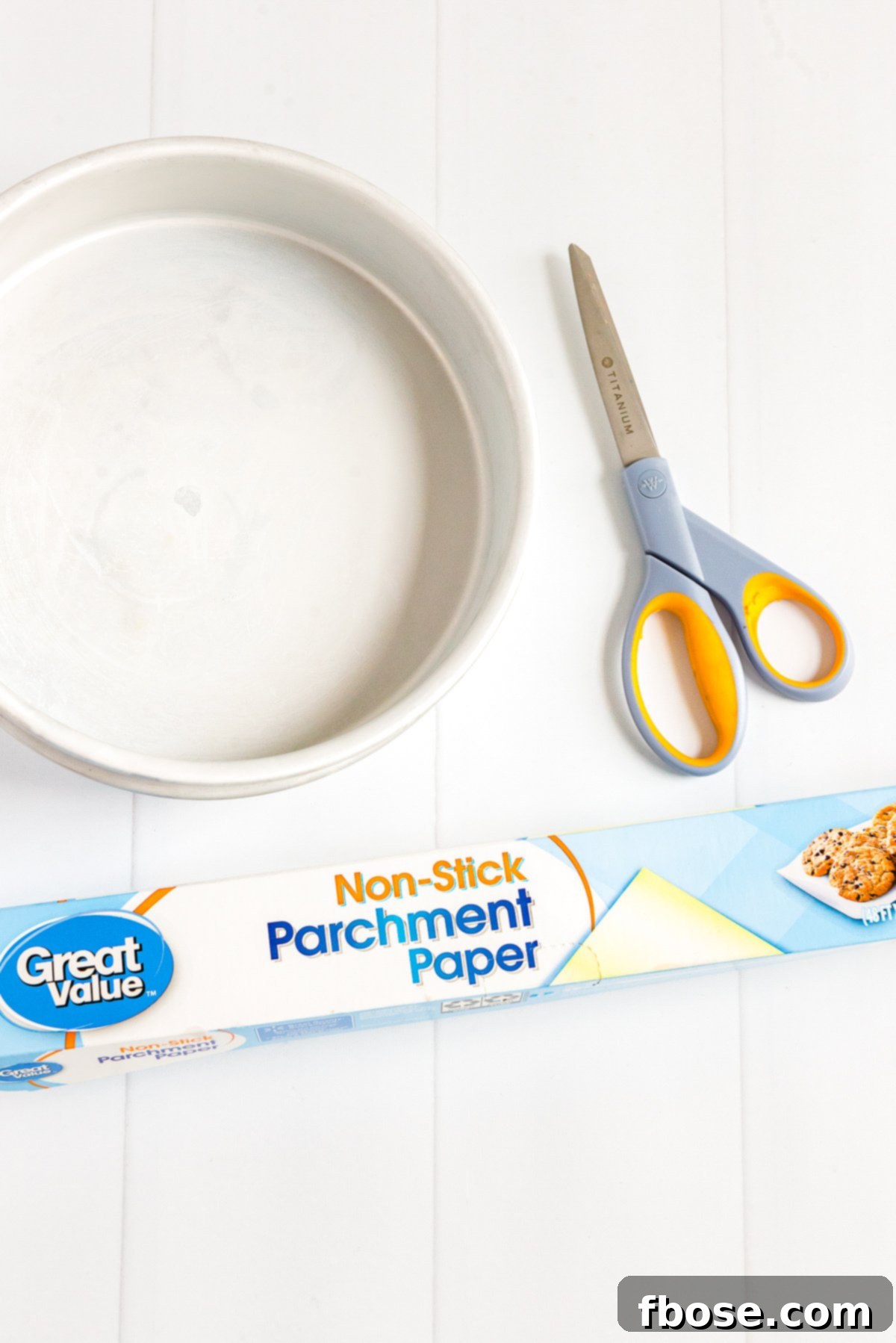 Cake pan, roll of parchment paper, and pair of scissors sitting on a white wooden table, ready for baking preparation.
