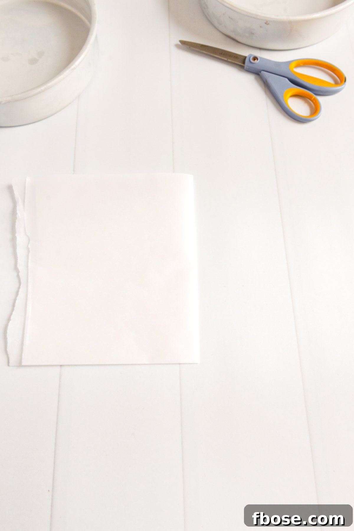 Piece of parchment paper folding in half with cake pans and scissors on the table.