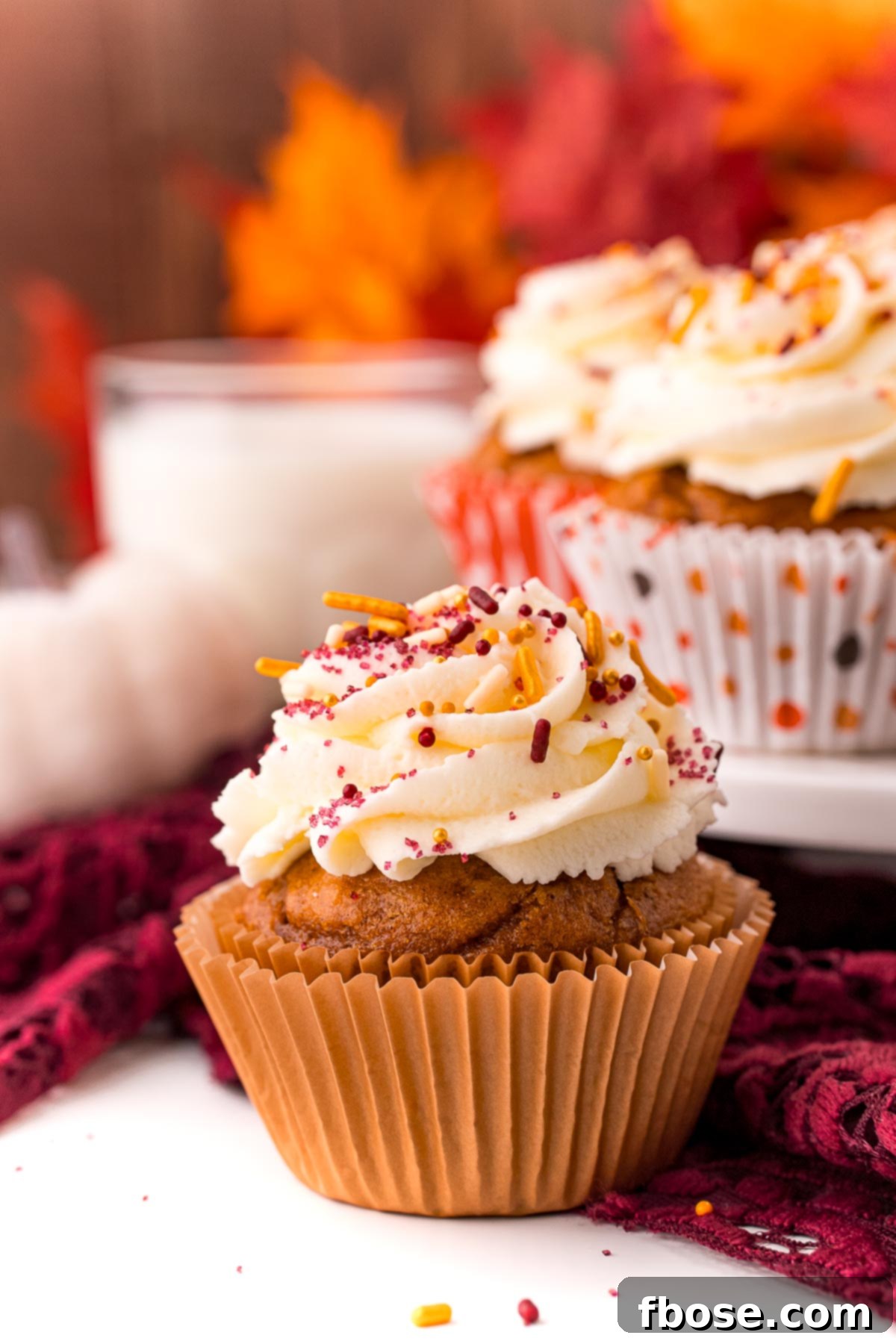 Close up photo of a pumpkin cupcake on a white table with a maroon napkin and more cupcakes and a glass of milk in the background. This image highlights the moist texture and the fluffy frosting, inviting viewers to imagine the delicious taste.