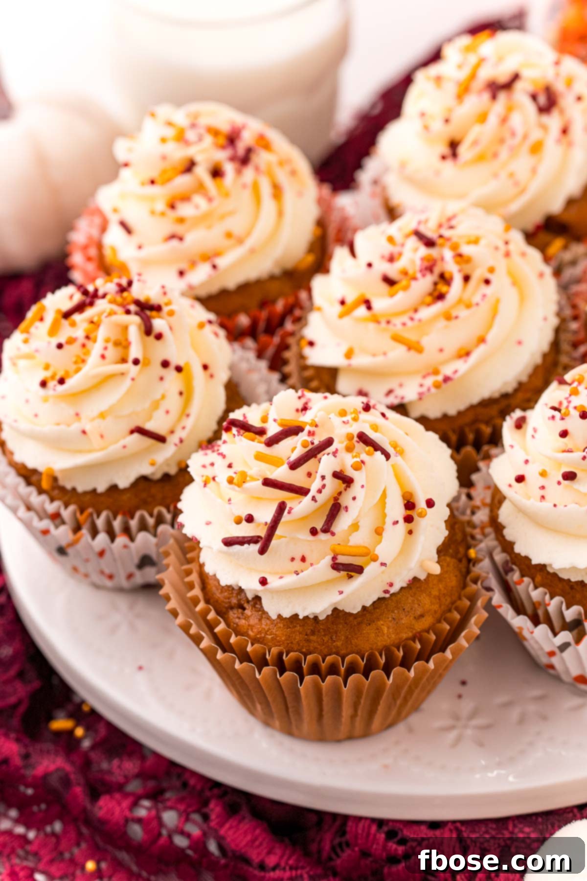 Pumpkin cupcakes on a white cake stand with a maroon napkin under it. The setting evokes a cozy fall atmosphere, emphasizing the seasonal appeal of the dessert.