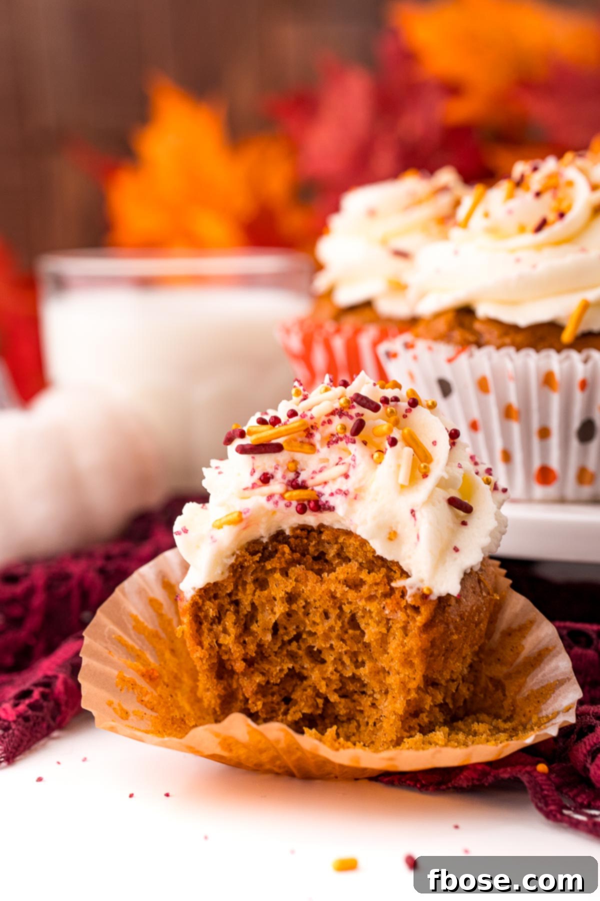 A pumpkin cupcake with a bite taken out of it on a white table with a cake stand with more cupcakes and a glass of milk in the background. This close-up shot emphasizes the deliciousness and homemade quality of the dessert.