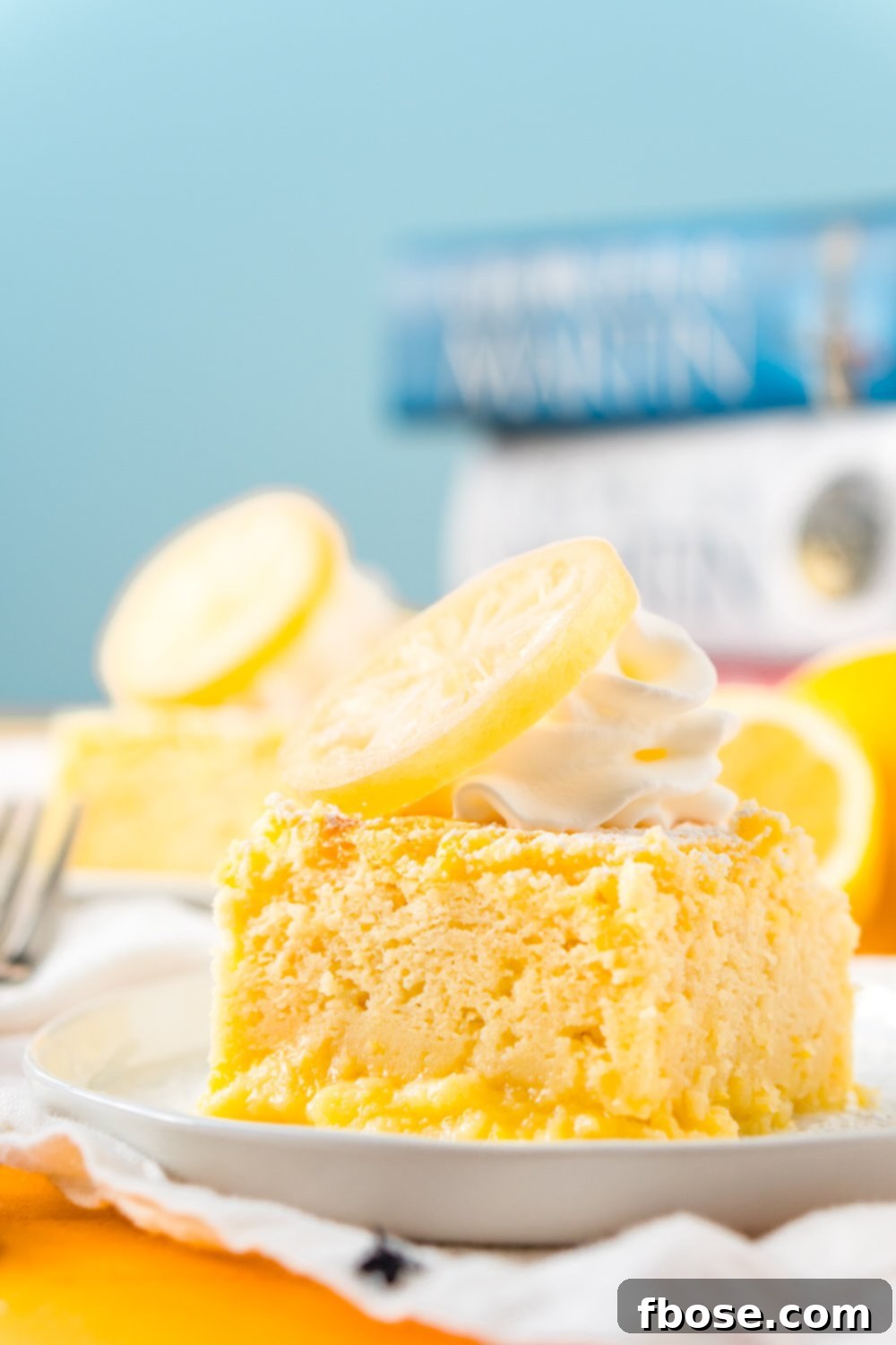 Piece of Lemon Magic Cake on a white plate with another slice of cake in the background and a fork next to it on the table.