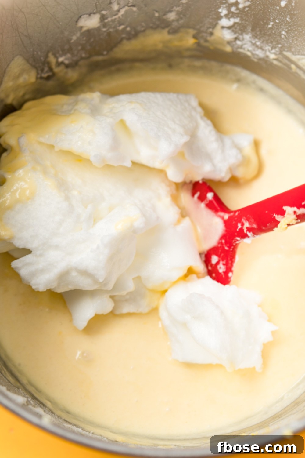 Stiff egg whites being folded into lemon cake batter in a metal mixing bowl.