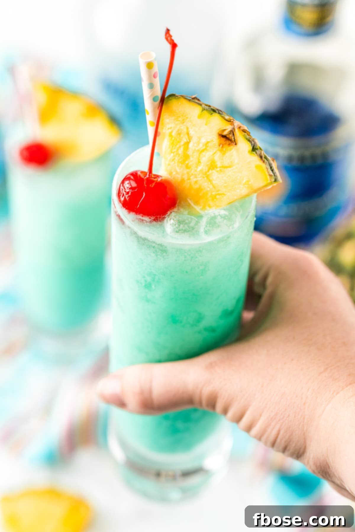 Azure Aloha 5 Woman's hand holding a blue hawaiian cocktail garnished with pineapple wedges and a maraschino cherry.