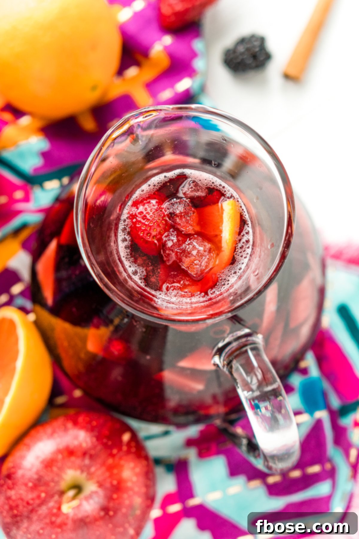 An inviting overhead shot of a pitcher brimming with vibrant red sangria, showcasing a colorful medley of fresh apples, blackberries, oranges, and strawberries.