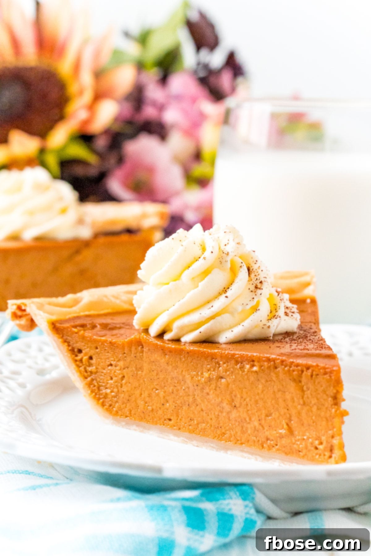 The Ultimate Pumpkin Pie 3 A beautifully baked slice of pumpkin pie on a white plate, accompanied by a glass of milk and autumnal flowers in the soft background.