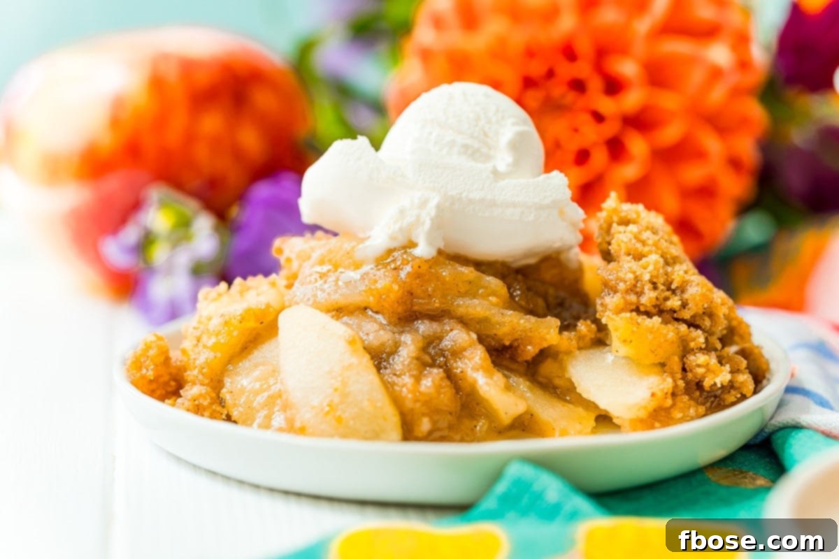 Photo of a serving of warm apple brown betty on a white plate, garnished with mint and fresh berries, with colorful flowers in the background.