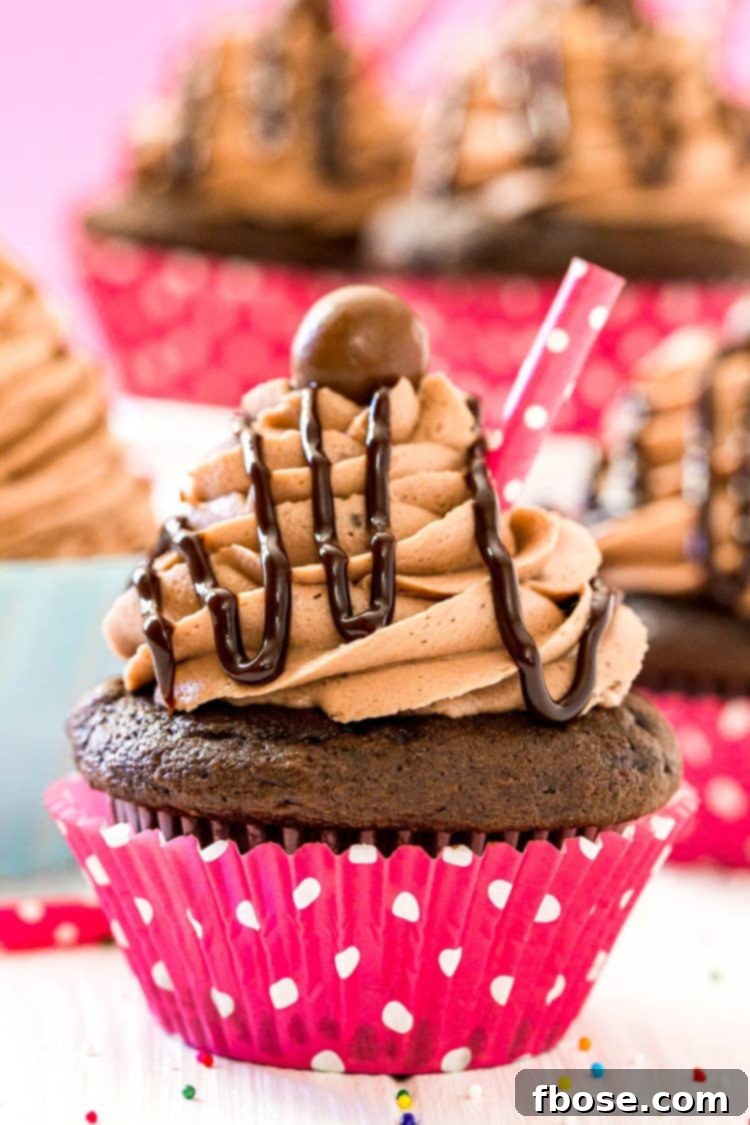 A detailed close-up of a single, perfectly crafted chocolate milkshake cupcake, adorned with creamy frosting, a chocolate drizzle, and a mini paper straw, with more delightful cupcakes blurred in the background.