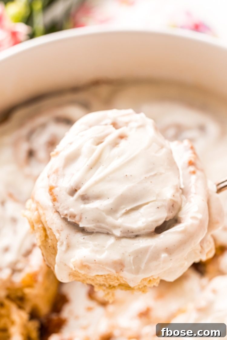A freshly baked cinnamon roll covered in creamy vanilla icing being carefully lifted from the pan, showcasing its golden-brown exterior and generous swirl of cinnamon filling.