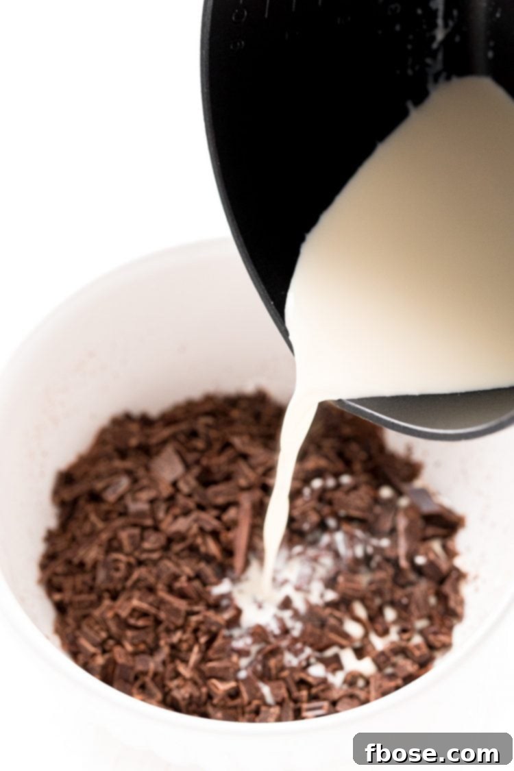 Hot heavy cream being carefully poured from a small saucepan into a white bowl filled with chopped chocolate.